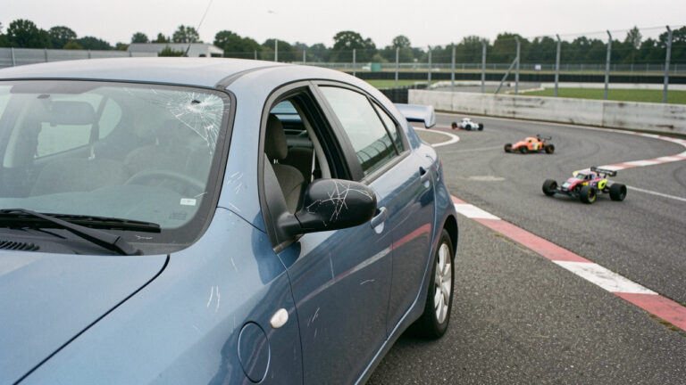 Voiture garée près d’un circuit de voitures télécommandées, présentant de légers dégâts de vandalisme comme des rayures et un rétroviseur fissuré.